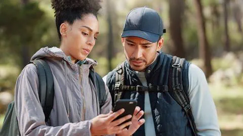 Two hikers, a woman and a man, examine the Blue Mountain App together in a wooded area, both wearing backpacks and outdoor gear.