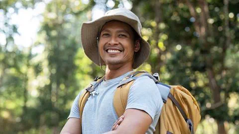 A smiling man with a hat and backpack outdoors in a sunlit forest.