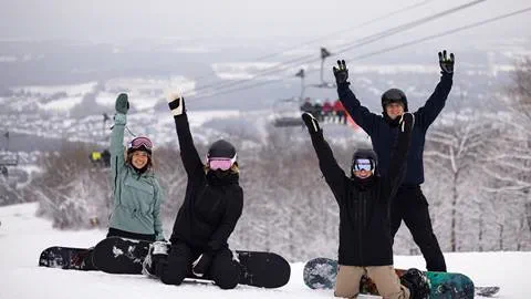 Group of snowboarders posing happily on a snowy mountain slope with a ski lift in the background.