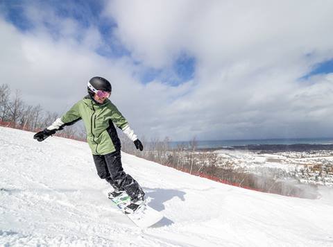 Person in a green jacket and black helmet snowboarding on a snowy slope with a panoramic view of a town and water in the background.