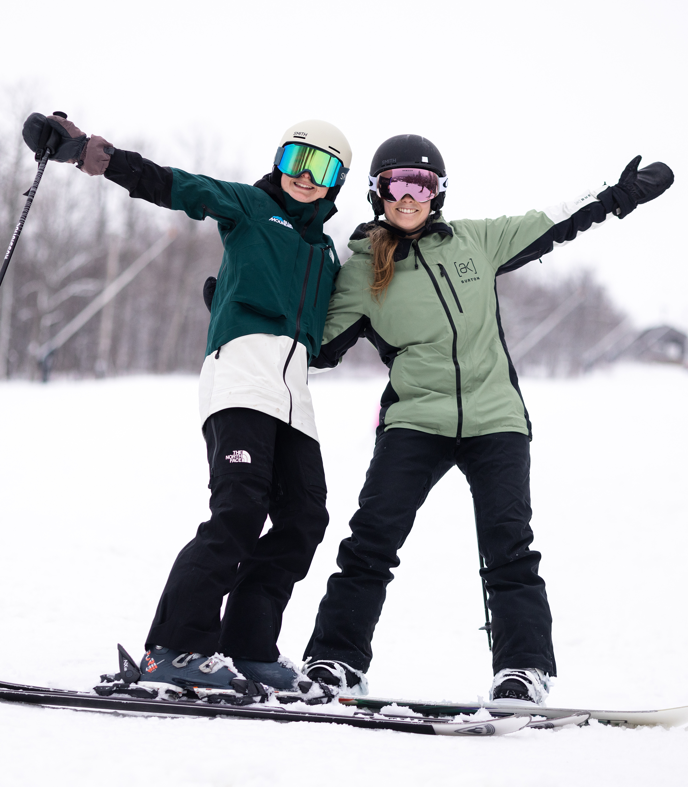 Two people in ski gear, including helmets and goggles, are sitting on a ski lift together, smiling at the camera. Snow-covered ground and bare trees are visible in the background.
