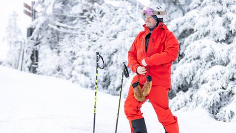 Person in bright red ski outfit stands in snowy landscape holding ski poles, surrounded by snow-covered trees.