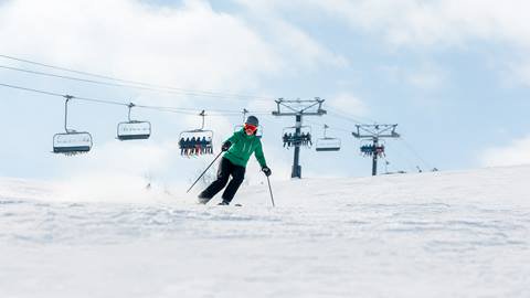 A skier in a green jacket and red goggles skis downhill on a snowy slope with ski lifts and people in the background under a cloudy sky.