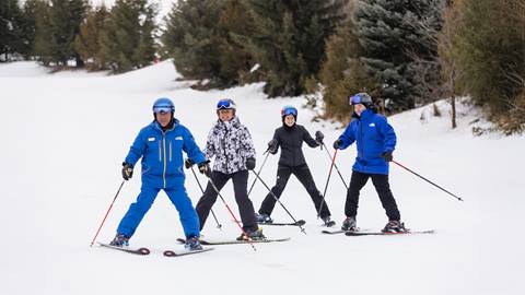 Four people skiing on a snow-covered slope, wearing blue and black ski gear, with trees in the background.