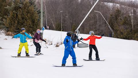 Four people snowboarding down a snowy slope, wearing colorful winter gear and helmets, with trees and a ski pole in the background.