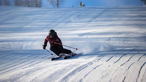 A person in a helmet and skiing gear is skiing downhill on a snowy slope, creating a trail of snow behind them under a clear blue sky.