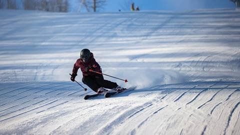 A person in a helmet and skiing gear is skiing downhill on a snowy slope, creating a trail of snow behind them under a clear blue sky.