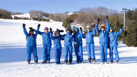 Eight people in matching blue ski outfits stand on the snow with their arms raised, trees and a snowy hill in the background.