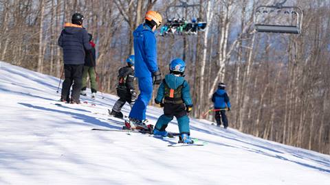 Several people, including children in helmets, skiing downhill with trees and a ski lift in the background.