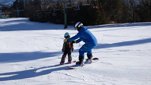 An adult in blue ski gear is teaching a child in colorful attire to snowboard on a snowy slope. They are both wearing helmets and goggles. Ski lifts and trees are visible in the background.