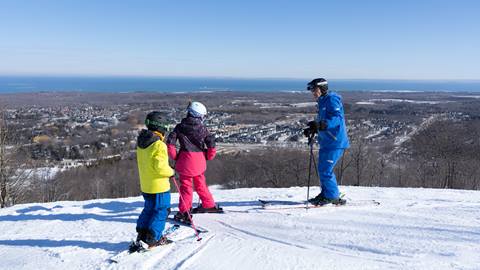 Three skiers, two children in bright outfits and an adult in blue, stand on a snowy slope overlooking a town and distant water under a clear sky.