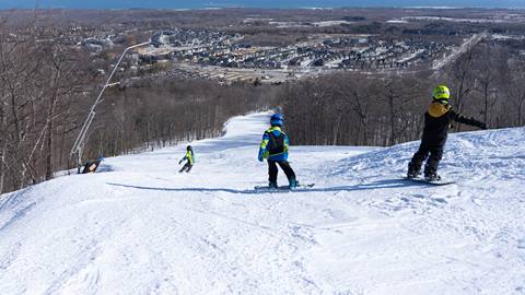 Skiers descend a snow-covered hill with a view of a town and tree-lined slopes in the background.