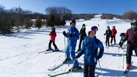 A group of skiers, including children and adults, stand on a snowy slope. They are dressed in colorful winter gear, each holding ski poles, with a clear blue sky above.