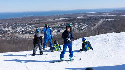 A group of children and an adult are skiing on a snowy slope with a town and a body of water visible in the background.
