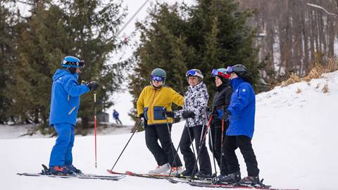 A ski instructor in blue talks to a group of four skiers on a snowy slope, with trees in the background.