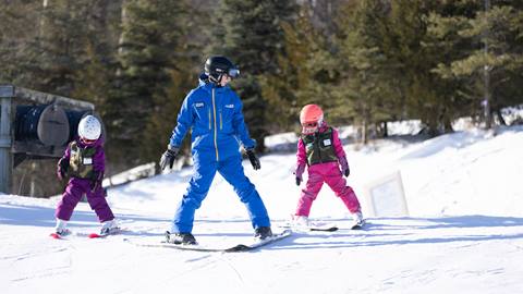 An adult in a blue ski suit instructs two children in pink ski outfits on a snowy slope, with trees in the background.