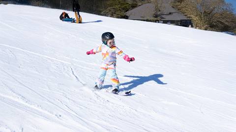 Child skiing down a snowy slope, wearing a colorful outfit and helmet. A person is visible in the background seated on the snow. Trees and a building are in the distance.