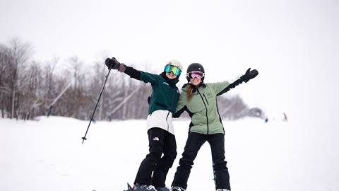 Two people are skiing on a snowy slope, posing with arms raised. They are wearing helmets, goggles, and winter gear. Trees in the background.