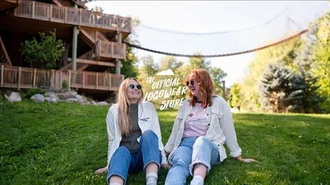Two women sitting on grass and smiling, with an overlay text "the official logowear store!" and a suspended bridge in the background.