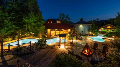 Outdoor pool area at night with illuminated swimming pools, a fire pit surrounded by chairs, and buildings in the background, set against a wooded landscape.