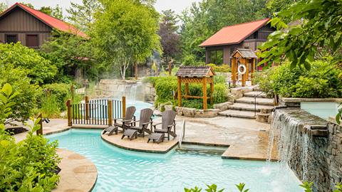 Scenic outdoor spa area with pools, lounge chairs, stone paths, lush greenery, and a small waterfall.