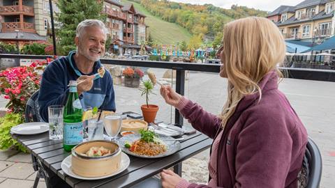 Two individuals enjoying an outdoor meal at a restaurant with a scenic village background.