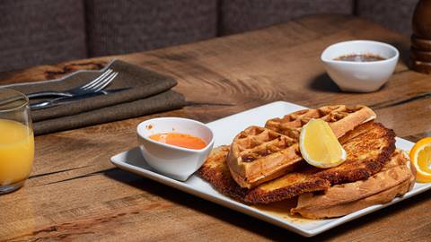 A plate of waffles with lemon wedges and a breaded fried filet, accompanied by syrup and a glass of orange juice on a wooden table.