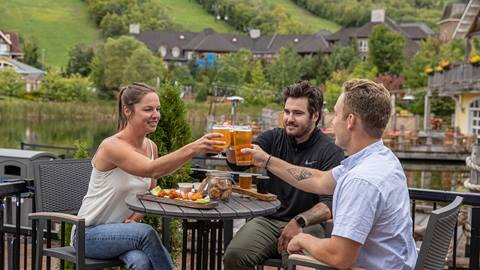 Three friends enjoying drinks and food outdoors with a scenic village backdrop.