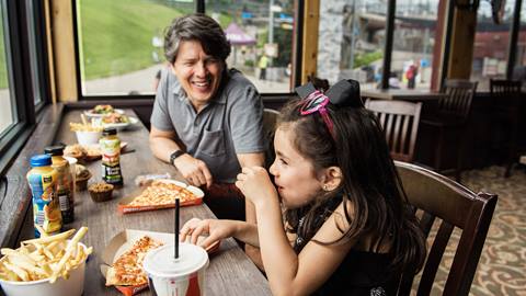 Father and daughter laughing and enjoying pizza and fries at a restaurant table by a window overlooking a grassy area.