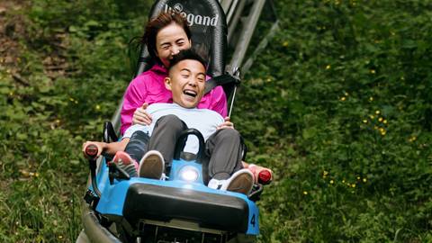 A joyful woman pushing a young boy in a motorized wheelchair through a grassy area, both smiling broadly.