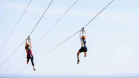 Two people zip-lining against a clear blue sky, equipped with safety gear including helmets and harnesses.