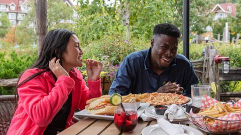 Two people laughing and enjoying a meal with burgers, fries, and pizza at an outdoor restaurant table.