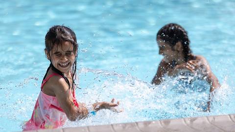 A young girl in a pink swimsuit laughs while splashing water in a pool, with another child partially visible in the background.