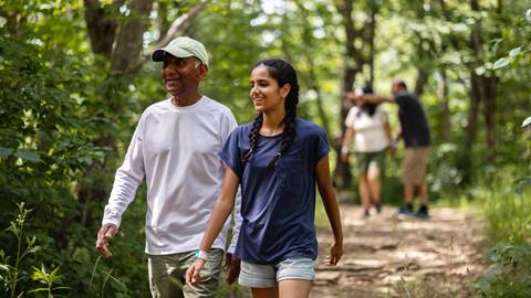 An older man and a young woman walking and talking on a wooded trail, with other walkers in the background.