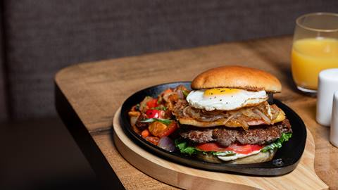 A gourmet burger with a fried egg, caramelized onions, and vegetables on a skillet, served with a glass of orange juice.