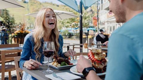 A woman laughing while dining outdoors with a man, with wine and a variety of dishes on the table.