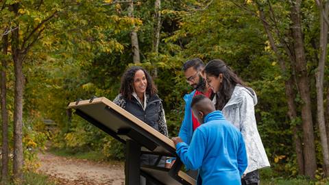 A diverse group of four people, including two adults and two children, are looking at an informational sign in a wooded park.