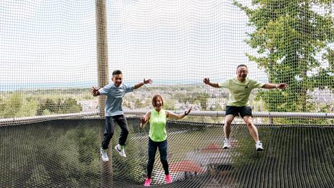 Three people joyfully jumping on a trampoline encased in a safety net, with a scenic landscape in the background.