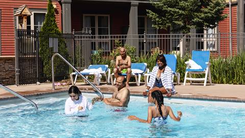 A diverse group of adults and children enjoying a sunny day in a swimming pool at a residential area, with lounge chairs and a building in the background.