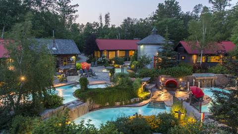 A luxurious outdoor poolscape with waterfalls, surrounded by lush greenery and lit buildings at twilight.