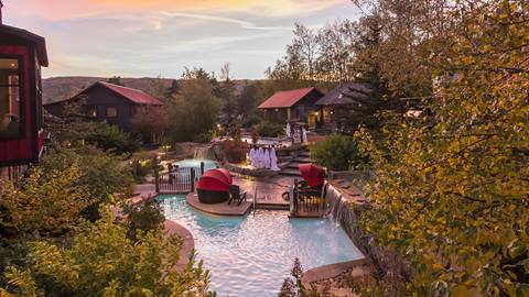 A tranquil, picturesque outdoor pool area surrounded by lush trees and rustic-style buildings, highlighted by a warm, sunset glow.