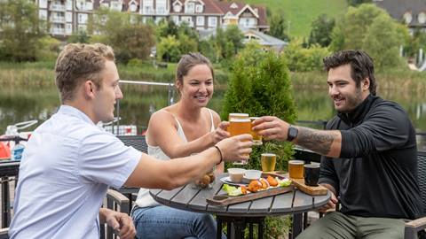 Three friends toast with beer at an outdoor table by a lake, with a backdrop of houses and green hills.