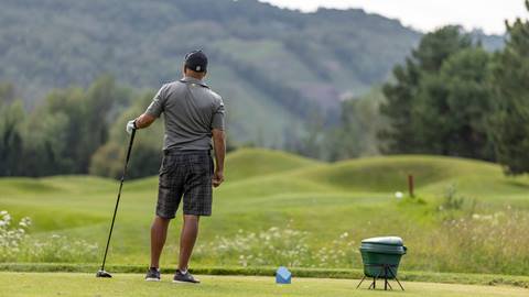 A golfer in plaid shorts and a gray shirt standing on a tee box, looking out over a green golf course with hills in the background.