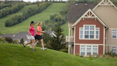Two women jogging on a grassy hill near large houses, wearing athletic gear.
