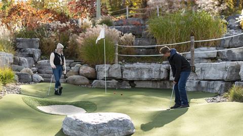 A man playing mini-golf focuses on his putt, while a woman watches, on a green surrounded by rocks and autumn foliage.