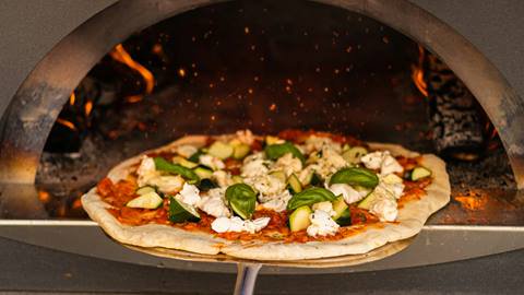 A pizza with basil and cheese toppings baking in a wood-fired oven, with visible flames on the side.