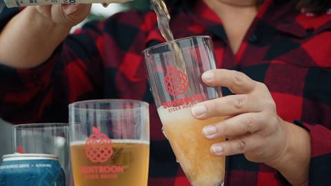 A person in a plaid shirt pours cider into a branded glass at an outdoor cider house.