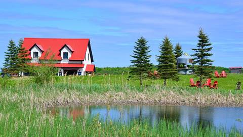 A picturesque rural landscape featuring a red-roofed house, surrounding greenery, and a small pond, under a clear blue sky.