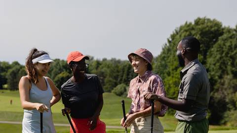 Four adults of diverse ethnic backgrounds laughing and holding golf clubs on a sunny golf course.
