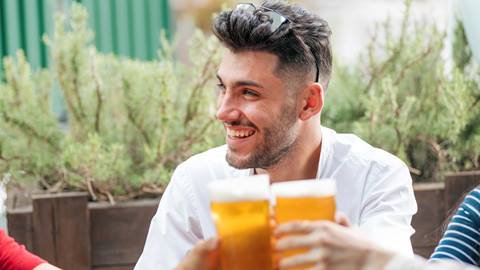 Trendy smiling man partying with his friends at an outdoor pub as they raise their glasses of cold beer in a toast with focus over the drinks to his face